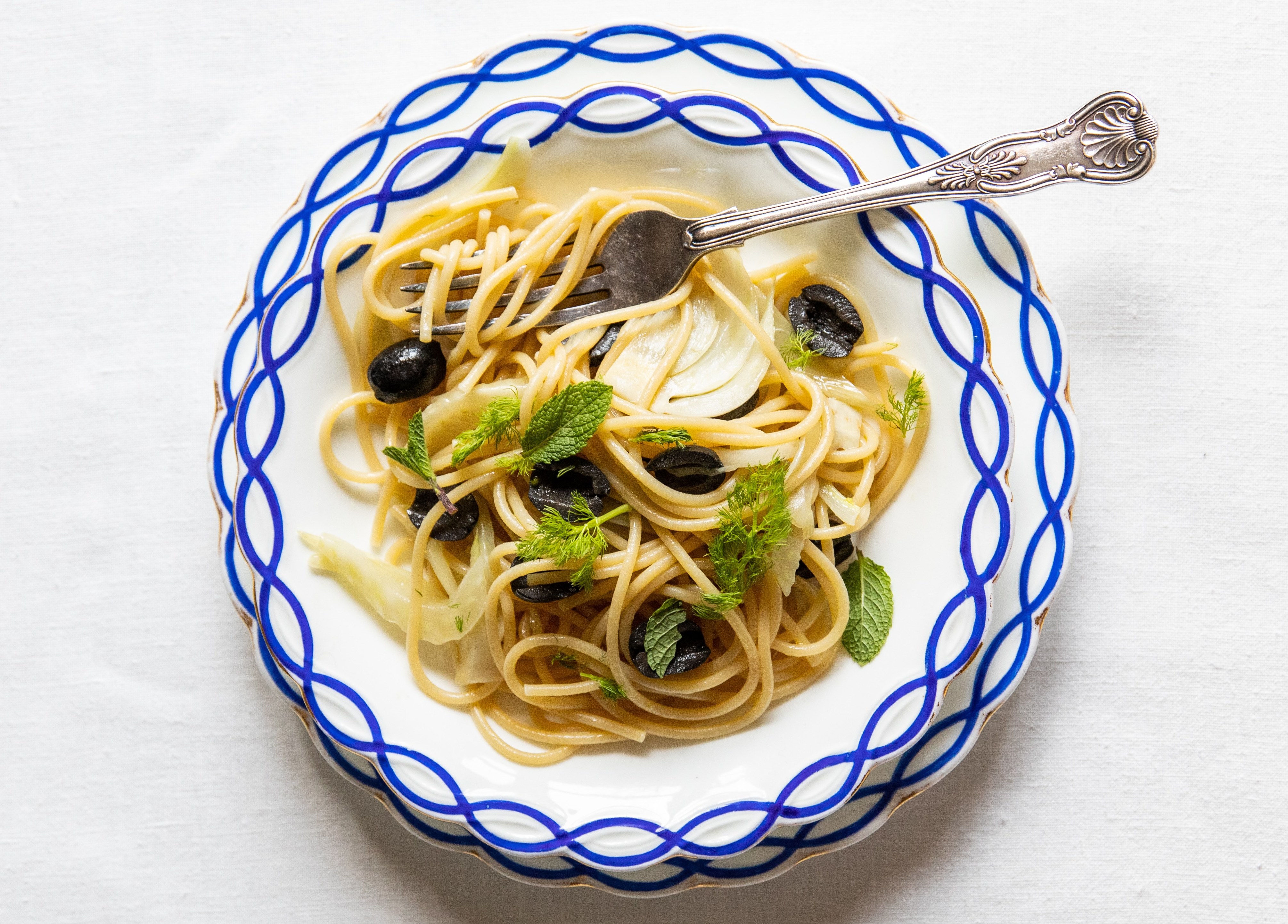 Spaghetti with olives and fennel on a blue and white plate.