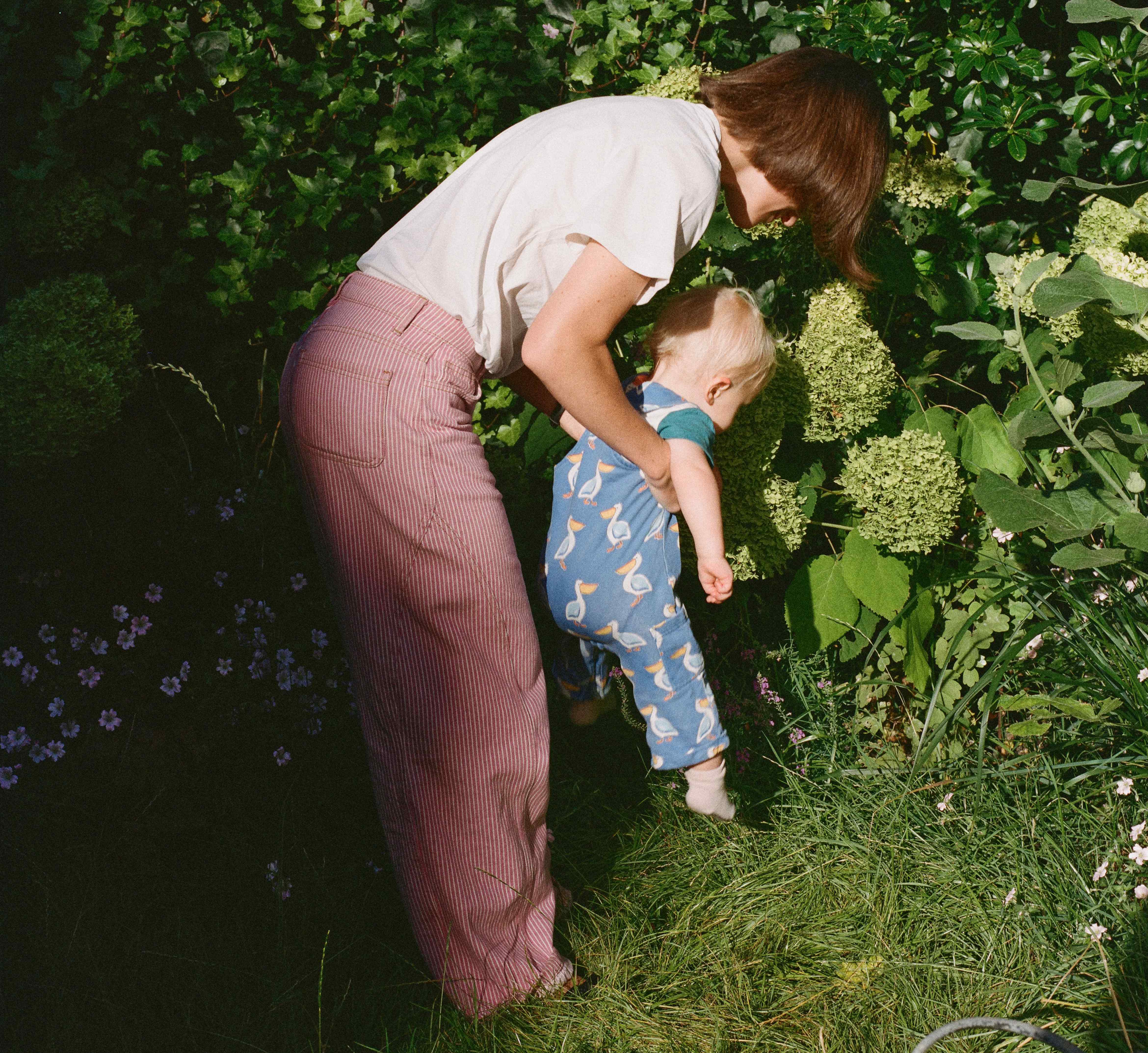 A woman helps a toddler walk in a garden.